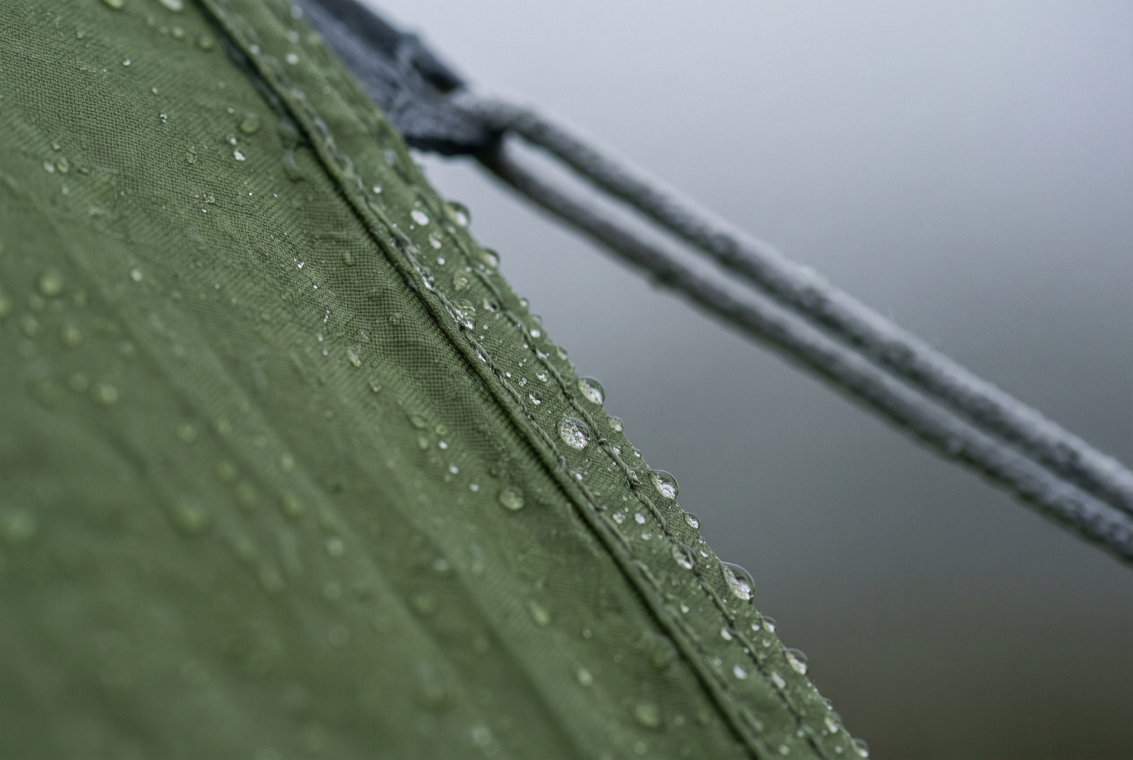 Rain running down taut tent fabric, drops caught along a seam with a guyline blurred behind