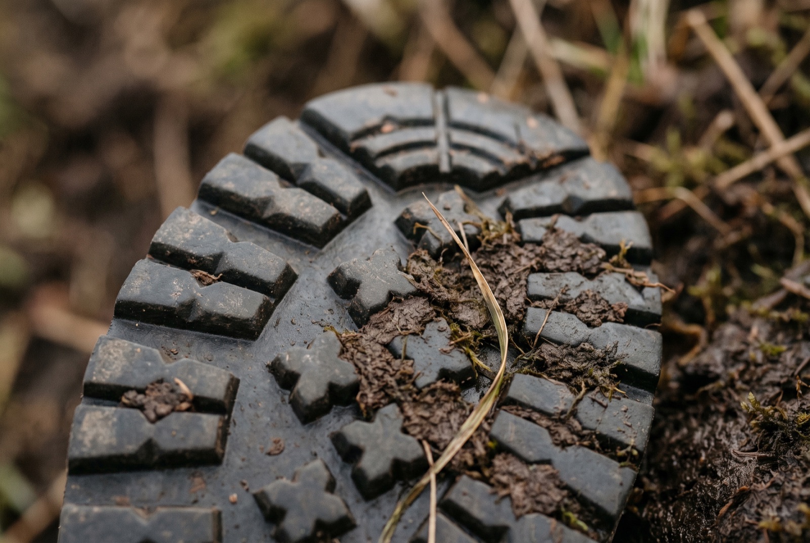 The underside of a hiking shoe pressed into wet peat, with clay and a blade of grass packed between the lugs