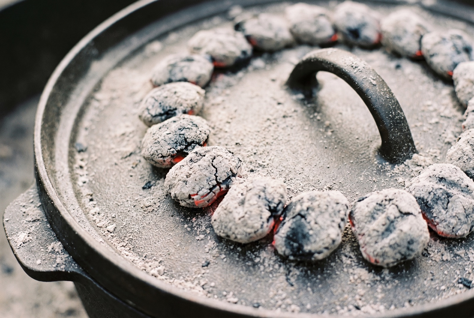 Greyed charcoal briquettes arranged across the flat lid of a camp dutch oven, some still glowing red at the edges.