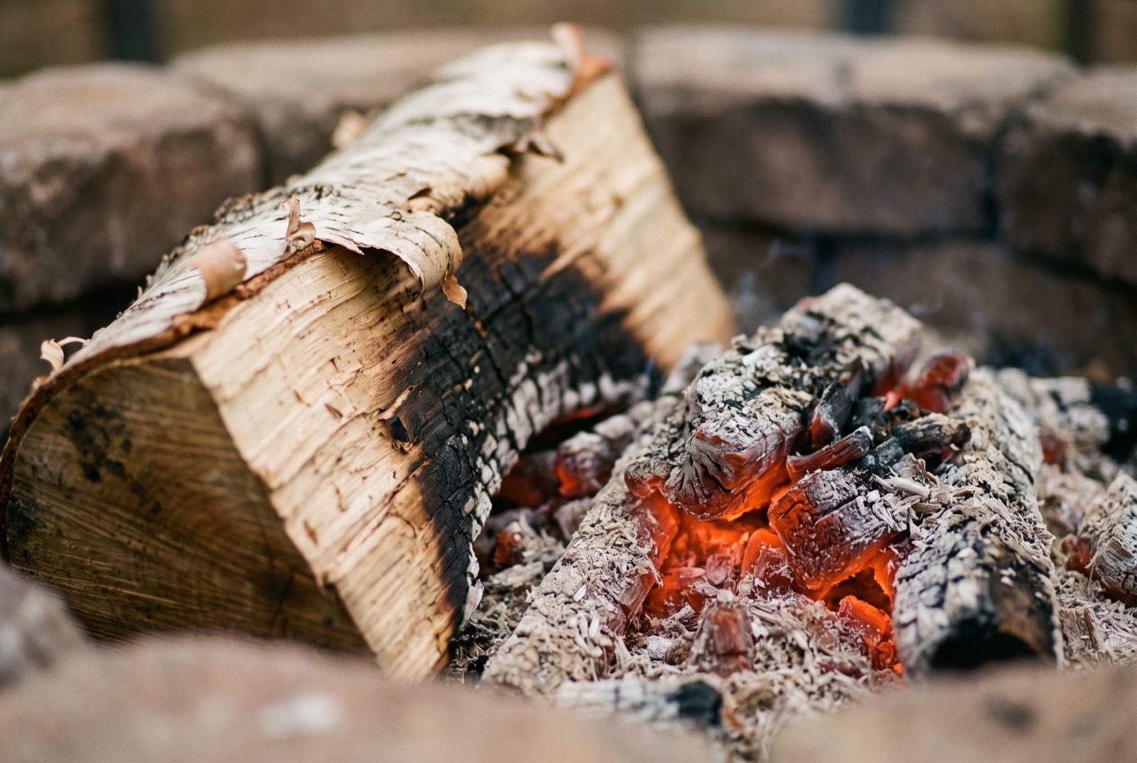 Glowing embers and a split birch log at the edge of a stone fire ring, pale ash against charred wood