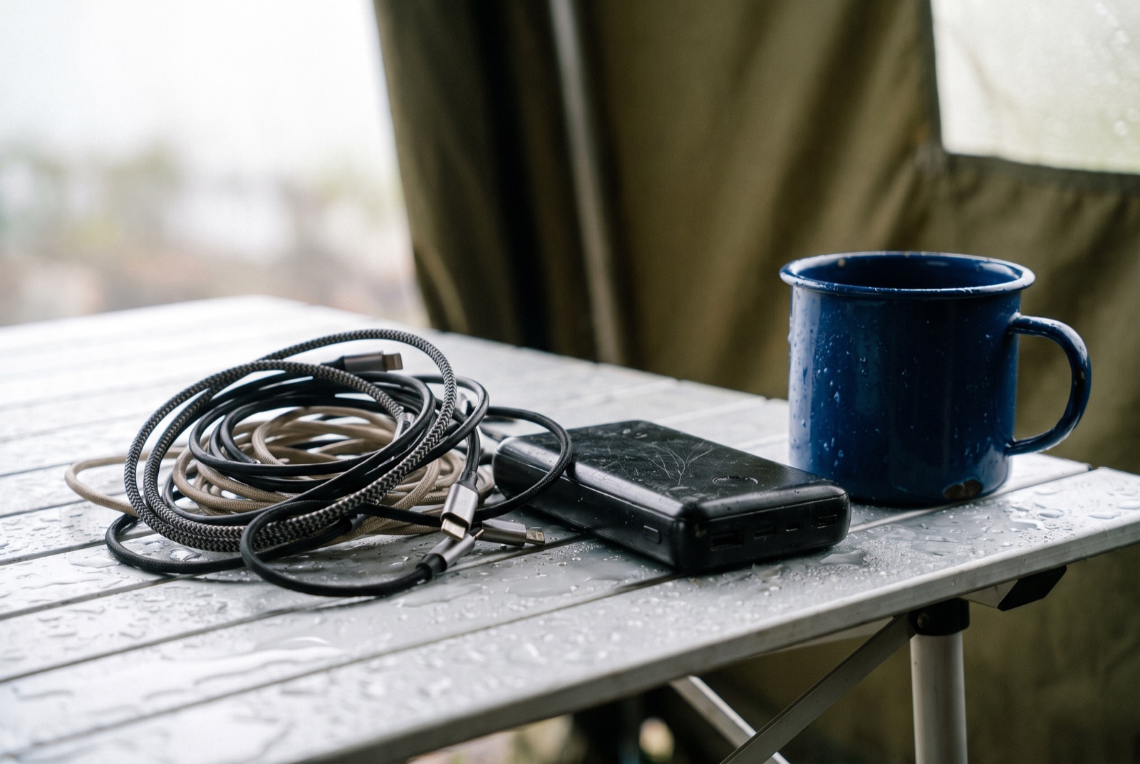 A worn power bank and a tangle of charging cables on a damp folding table beside an enamel mug