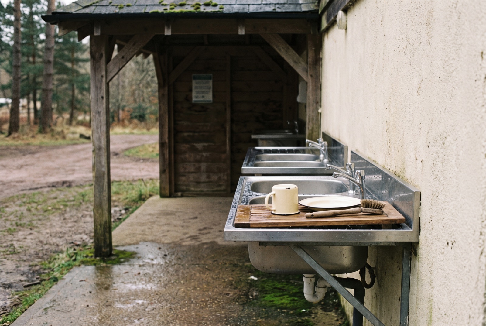 A row of stainless steel washing-up sinks under an open timber shelter at a campsite, with a single enamel mug and plate on the draining board.
