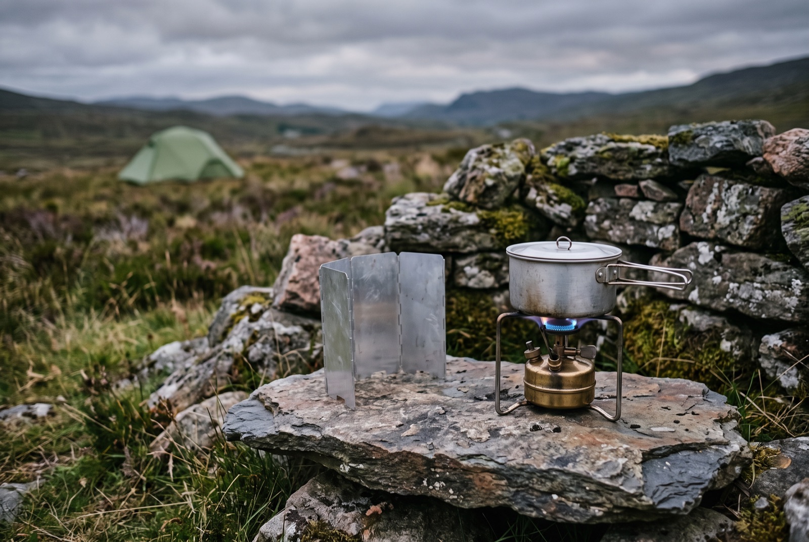 A camping stove burning in the open on a flat stone in the shelter of a drystone wall, with a windshield beside it and a tent in the distance.