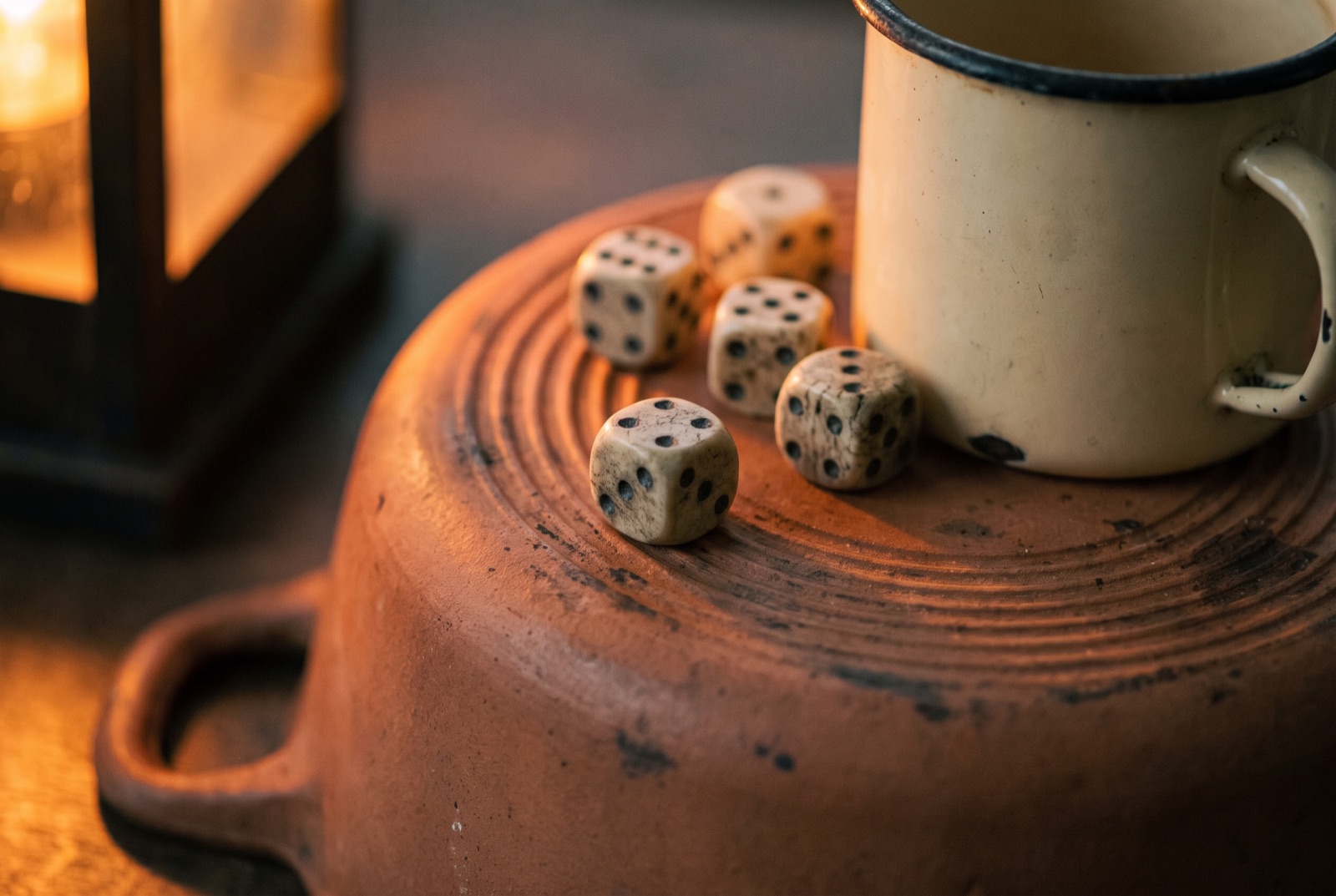 Five dice tipped from an enamel mug onto an upturned saucepan lid, lit from one side by a fire