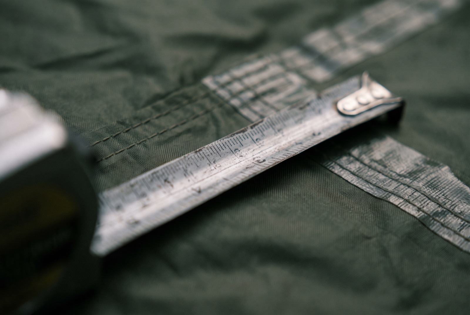 A tape measure extended across the corner of a tent groundsheet, the seam and fabric weave visible behind it