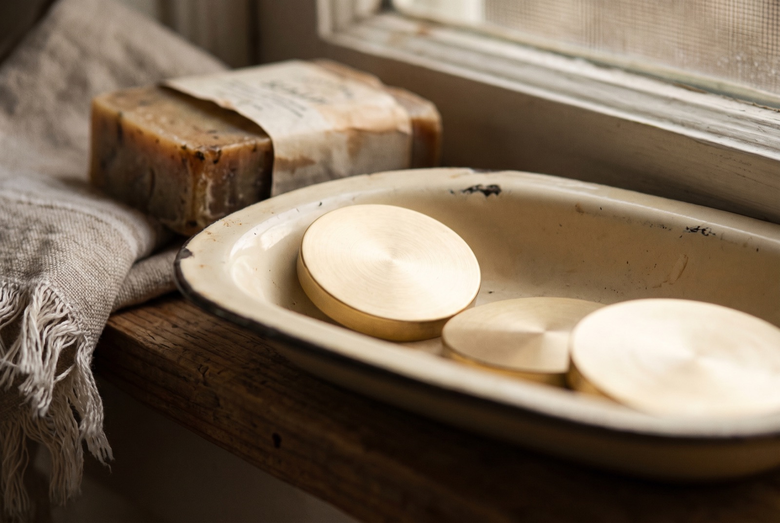 Blank brass shower tokens in a shallow enamel dish on a wooden shelf, beside a bar of soap and the edge of a folded towel.