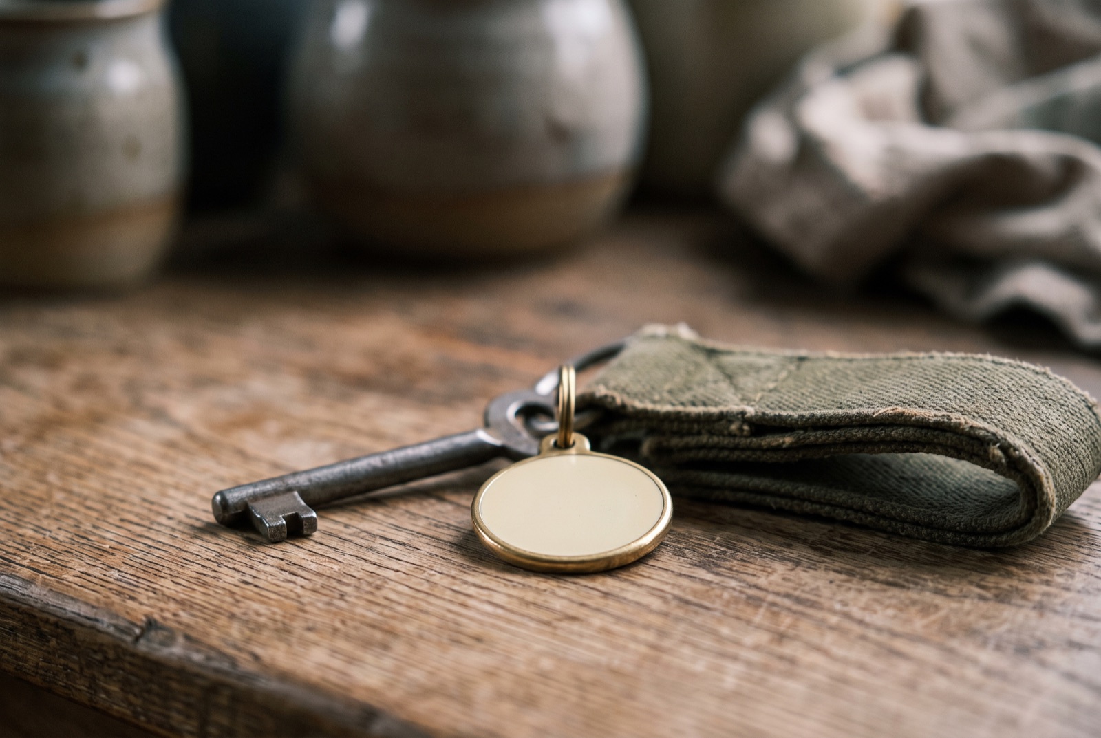 A plain iron key and blank metal fob on a scrubbed wooden counter beside a folded canvas strap.